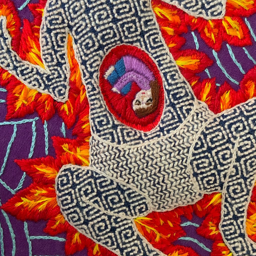 Close up of hand embroidery piece by Annie Davidson, featuring patterned figure outlined by fire with a human curled up in their stomach