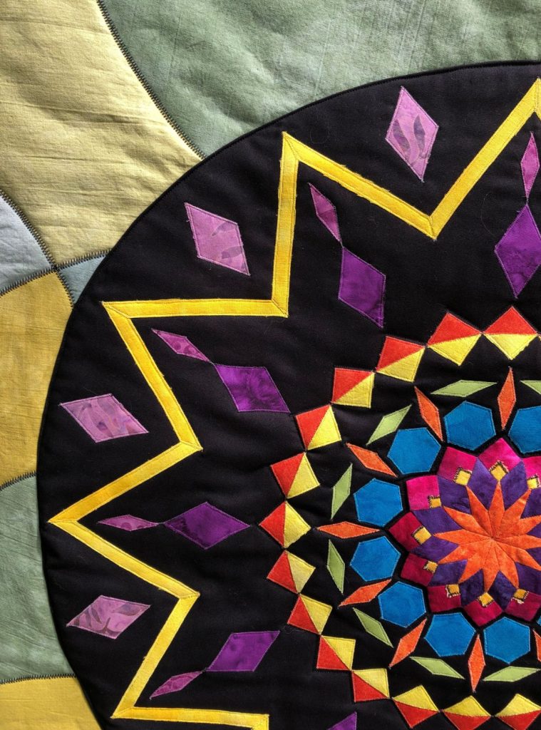 Close up of a patchwork quilting piece, featuring a black, gold and purple geometric design against a muted blue and green background